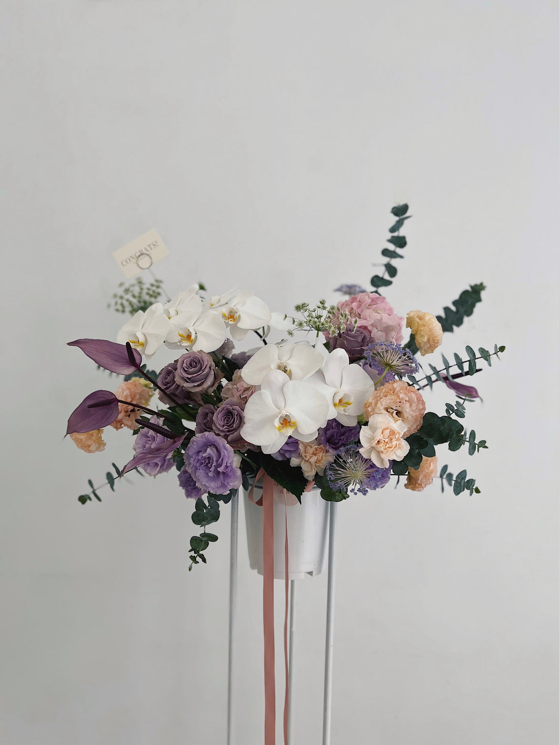 一束高架花籃,包含紫色玫瑰、白色蘭花、粉色和橙色康乃馨,以綠葉點綴。
