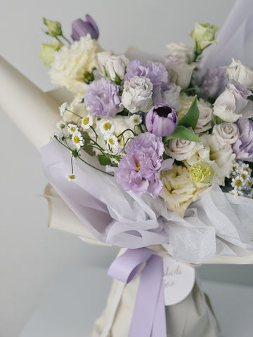 一束淡紫色和奶白色鮮花組成的花束,搭配小雛菊和綠葉,包裹在紫色絲帶中。