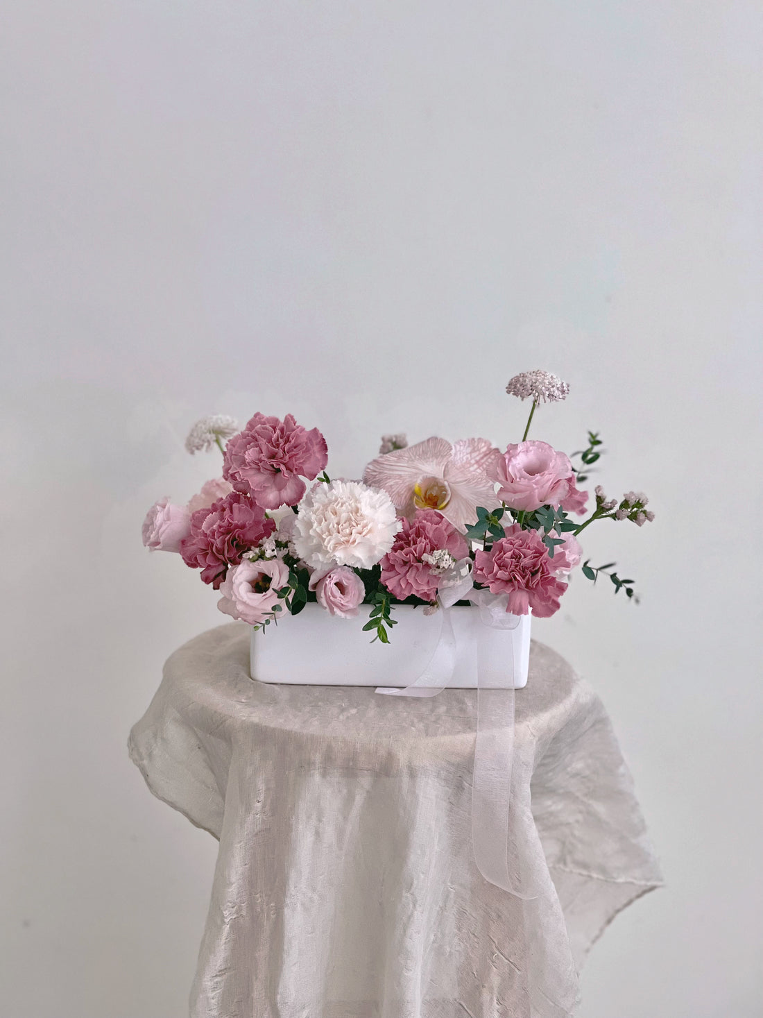 白色長方形花盆內,粉色和白色康乃馨組成的花束,置於淺色桌布上。
