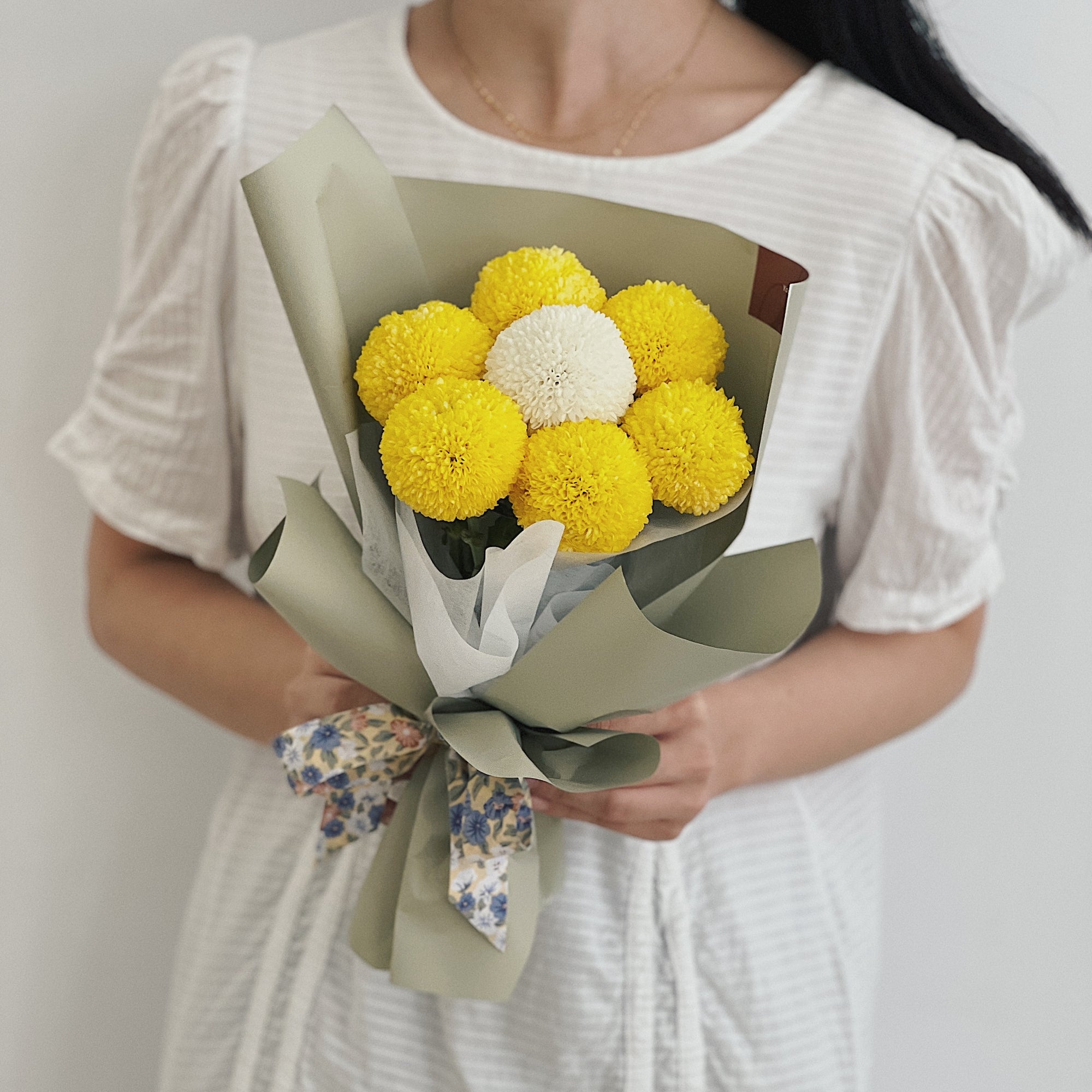 女性手持球型花束，花朵為黃色和白色，包裹於綠色包裝紙。