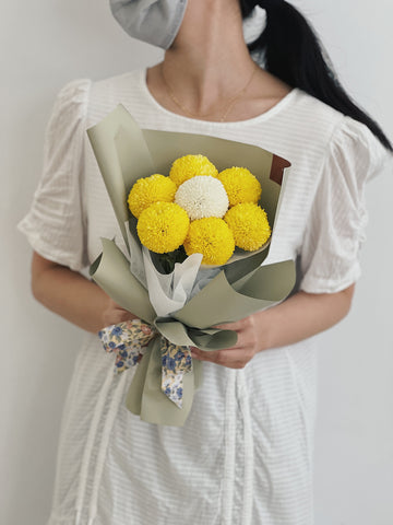 女性手持球型花束，花朵為黃色和白色，包裹於綠色包裝紙。