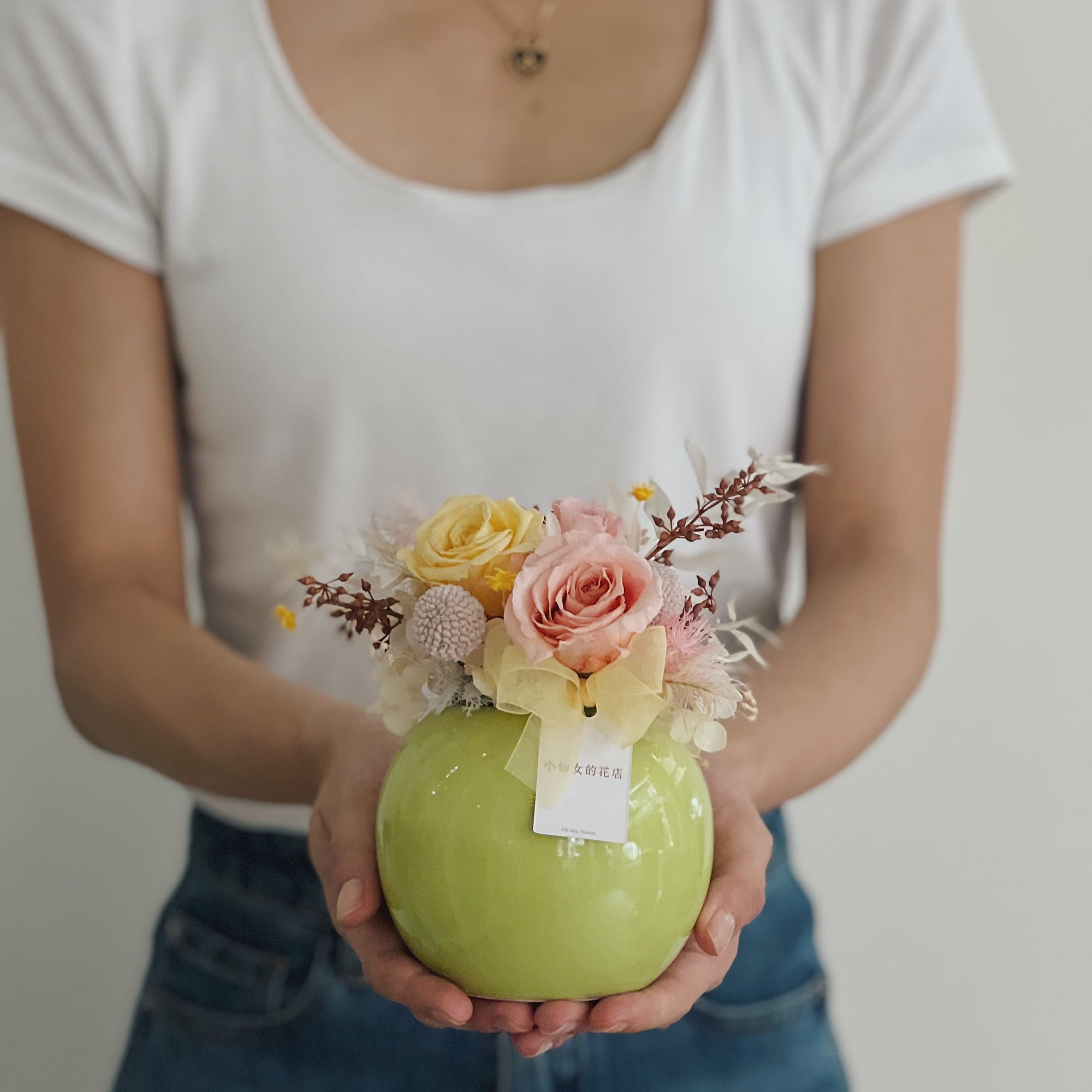 女人手持綠色圓形花盆，裡面有永生玫瑰和飾物。