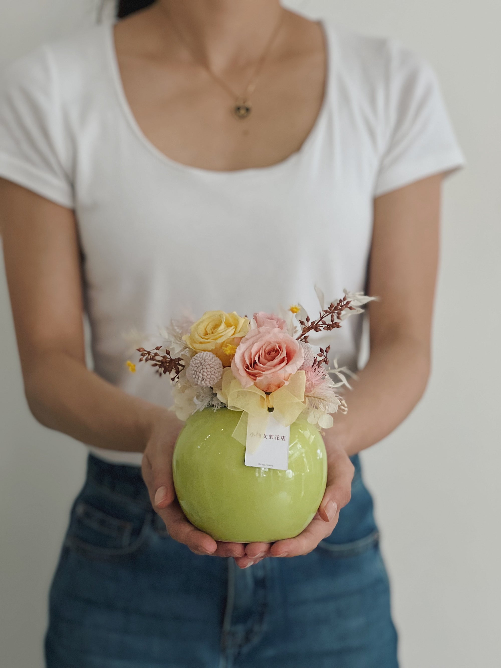 女人手持綠色圓形花盆，裡面有永生玫瑰和飾物。