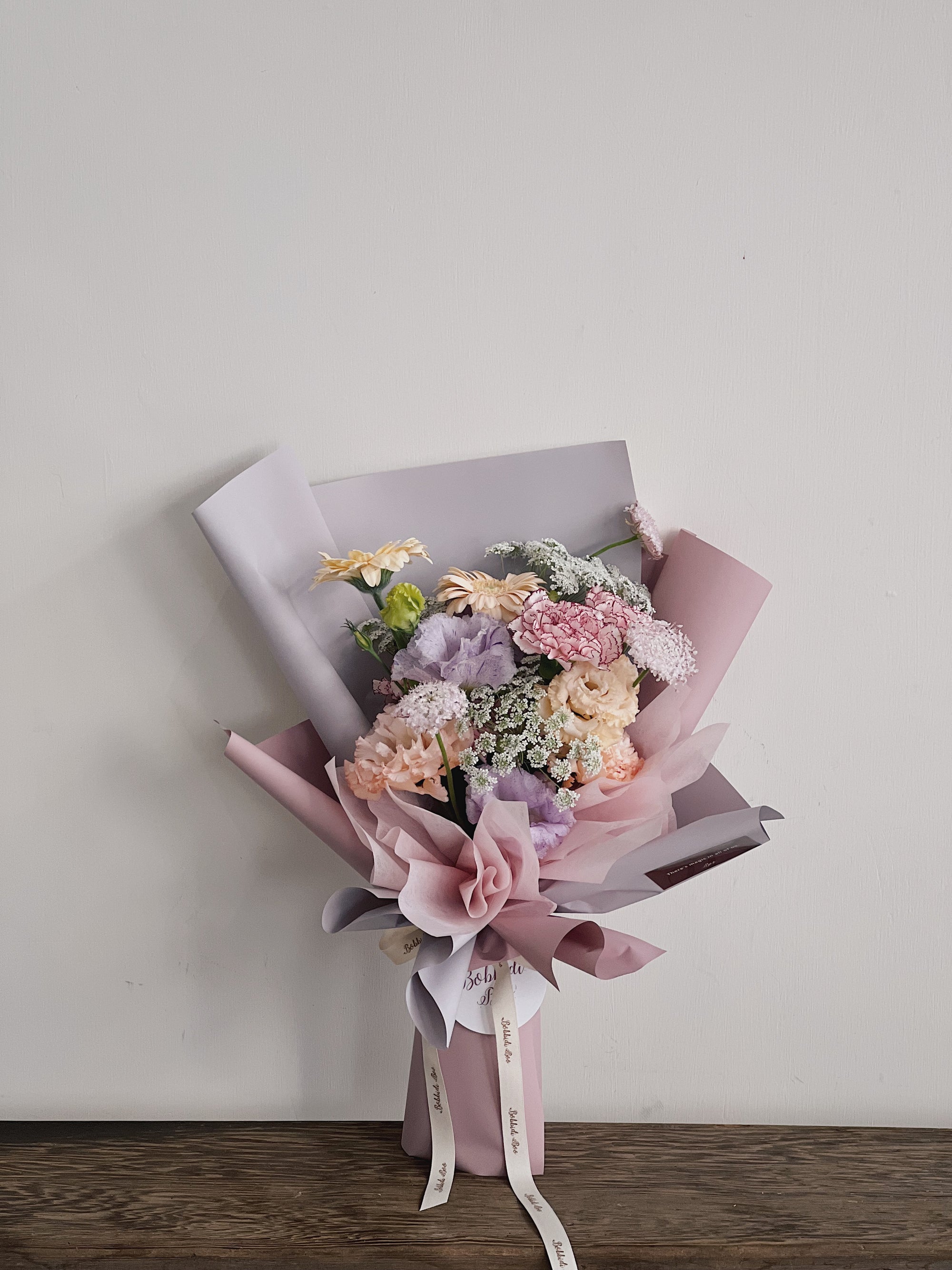 粉色和淡紫色的花束，包裹著多種鮮花，放在木桌上。