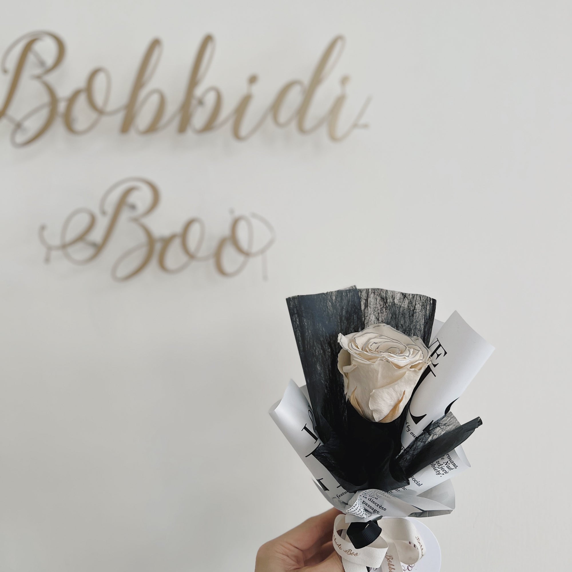 手握單枝包裝的永生玫瑰花，背景有「Bobbidi Boo」裝飾字。