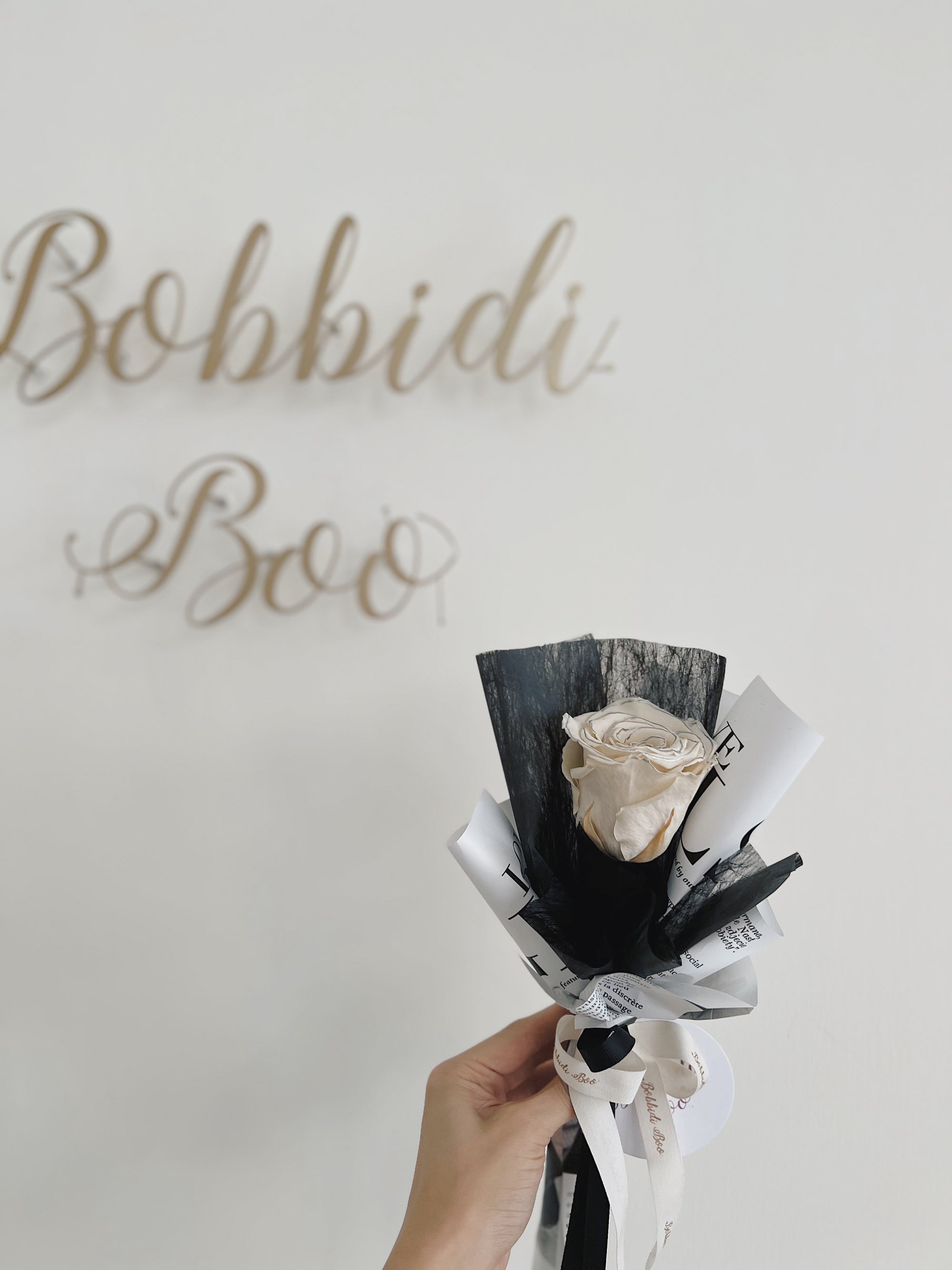 手握單枝包裝的永生玫瑰花，背景有「Bobbidi Boo」裝飾字。