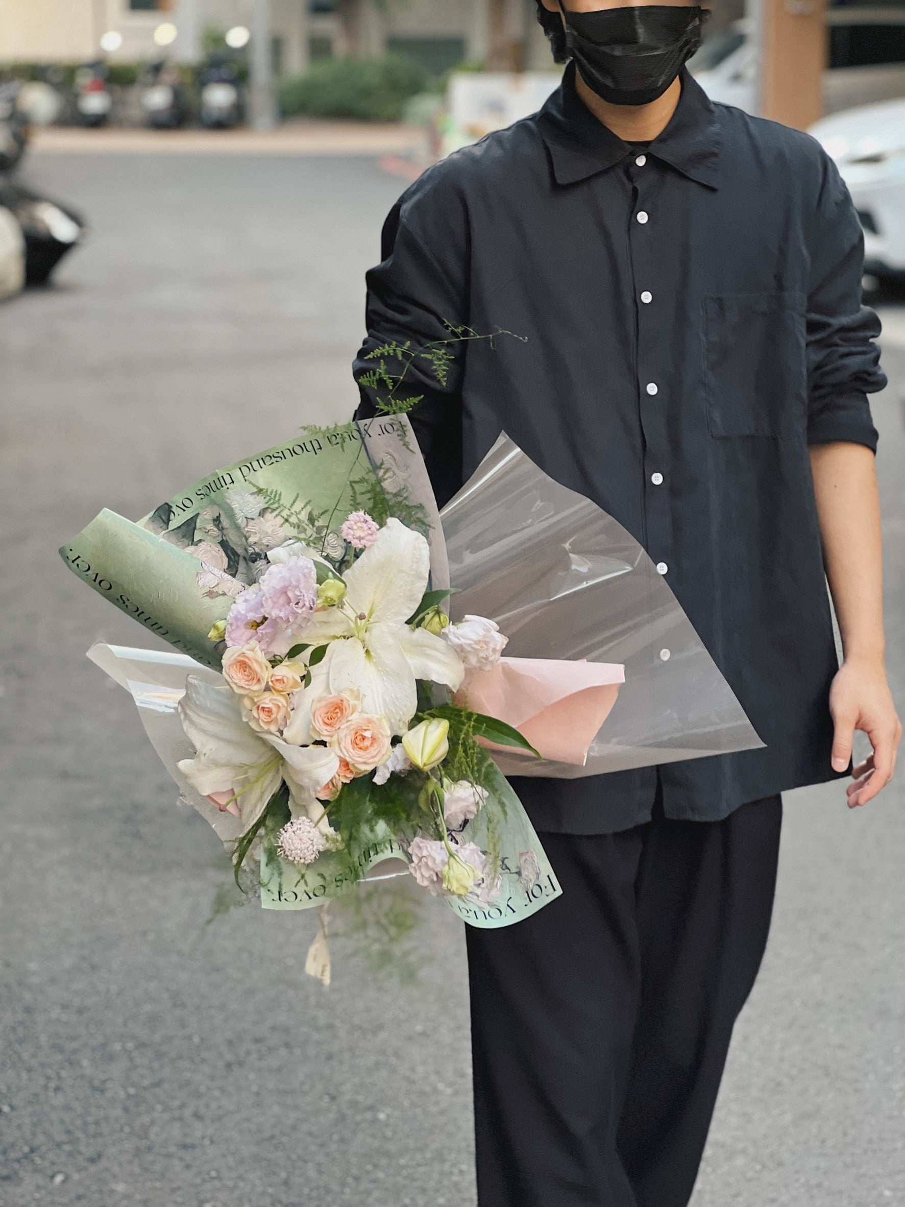 一名穿黑色襯衫和黑色口罩的人拿著綠色包裝的百合花花束。