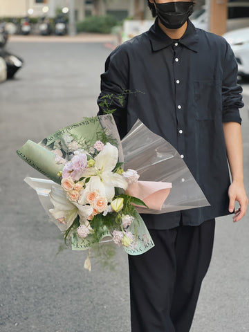 一名穿黑色襯衫和黑色口罩的人拿著綠色包裝的百合花花束。