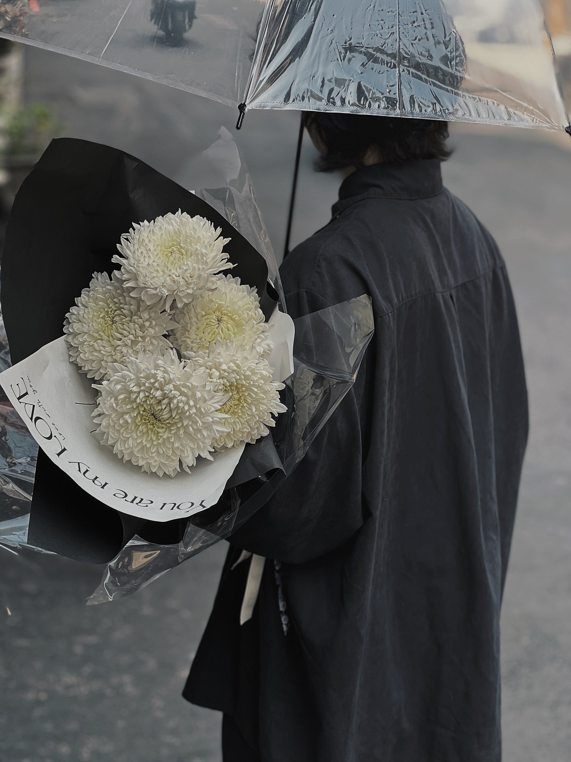 一人撐透明雨傘，手持白色菊花花束，背景為灰色街道。